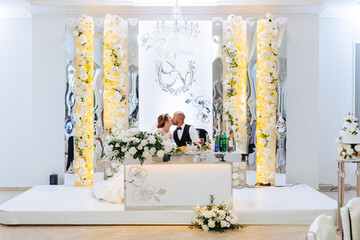 A bride and groom are kissing in front of a white table with flowers and a vase. The table is set for a wedding reception
