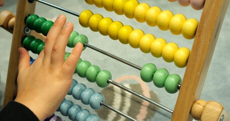 Little boy hand moves green balls on abacus counting in daycare classroom closeup. Toddler student learns to add numbers with colorful wooden toy