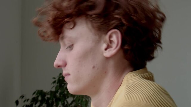 Side closeup of blue-eyed Caucasian man with ginger curly hair rotating head and stretching neck, relieving pain after sedentary work indoors