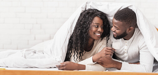 Safe sex. Young african american couple holding condom, lying on bed under white blanket, looking...