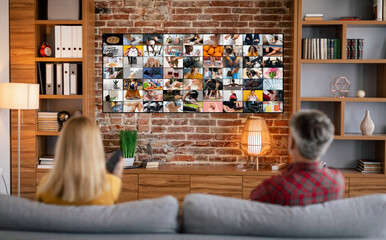 A couple relaxes on a couch in their living room, facing a large television screen displaying a grid of various images. The room features wooden shelves filled with books and decor, a brick wall