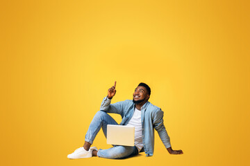 Great offer. Black millennial guy sitting on floor with laptop and pointing upwards at copy space on yellow background