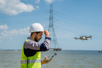 Obraz premium Engineer in a safety vest operating a drone to inspect and survey electrical power lines. Technician inspecting the power station for scheduled maintenance.