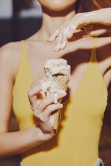 A young woman holding an ice cream cone with melting ice cream on her hands She is wearing a bright yellow tank top The background is softly blurred