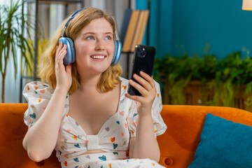 Redhead happy overjoyed woman in wireless headphones relaxing sitting on couch at home apartment choosing, listening favorite energetic disco dancing music. People weekend leisure activities