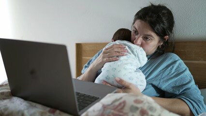 Mother working in front of the laptop while holding newborn in her arms, illustrating the balance...