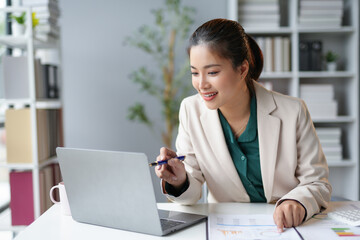Young asian businesswoman sitting at desk in modern office, analyzing financial charts on laptop and having video call
