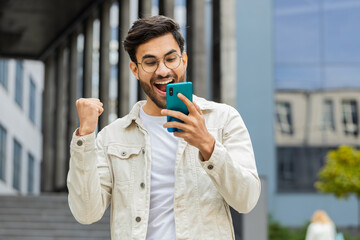 Young Indian man use mobile smartphone celebrating win good message news, lottery jackpot victory,...