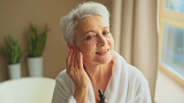 Woman applying natural drops cosmetics oil to face skin. Mature adult old woman applying nourishing anti age serum with pipette doing daily beauty routine. Senior woman gets kin scare facial treatment