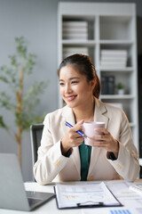 Smiling businesswoman holding a coffee cup and thinking about a business strategy while working on a laptop in an office