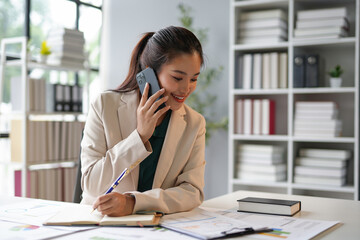 Young asian businesswoman sitting at desk in office talking on phone and taking notes in planner