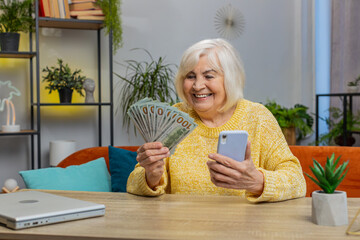 Planning family budget. Smiling excited senior woman counting money cash use smartphone calculate domestic bills at home. Old grandmother satisfied of income and saves money for planned vacation gifts