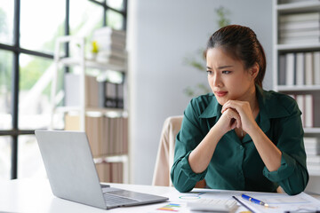 Young asian businesswoman is frowning and looking worried while working on a laptop in her office