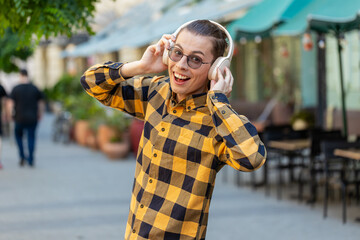 Fototapeta premium Rear view of happy relaxed man tourist in wireless headphones choosing, listening favorite energetic disco music on smartphone outdoors. Caucasian tourist walking passes by urban sunny city street.