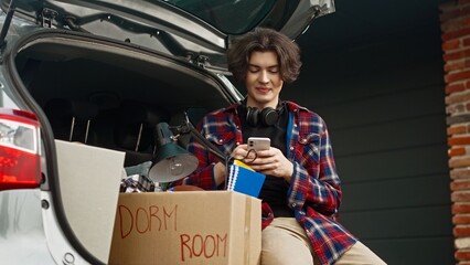 Young teenager sitting in auto trunk while chatting with his friends on smartphone. Remotely...