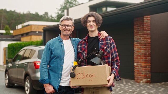 Portrait of father and his son standing outside in yard. Handsome dad hugging his child and proud of his good achievement in college. Satisfied people joyfully smiling while looking on camera.