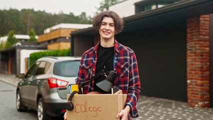 Portrait of Caucasian student holding box with his belongings while standing in yard. Young teen smiling with joy while looking at camera. Having good feelings before upcoming semester at college.