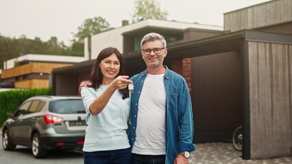 Happy Caucasian couple cuddling each other while standing before their new estate. Beautiful wife showing on camera keys to purchased house. Smiling with joy and happy about successful purchase.