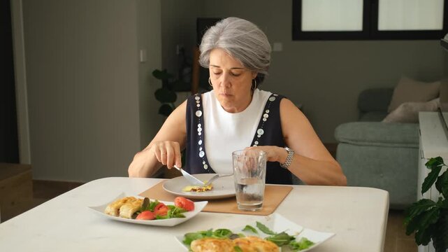 Senior woman eating a healthy, balanced meal alone at home, cutting food with a knife and fork. Salad, fruit, and bread are on the table in a cozy dining area