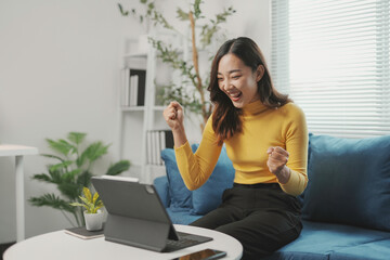 Young asian woman is raising her arms in celebration while looking at a tablet on the table