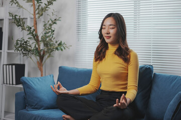 Young asian woman is practicing mindfulness with her eyes closed while sitting in the lotus position on a sofa at home