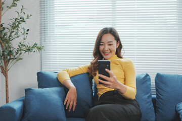 Young woman is sitting on a comfortable sofa, smiling while using her smartphone