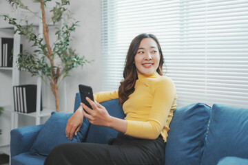 Obraz premium Young woman relaxing on a blue sofa is using a smartphone and smiling while looking away from the camera
