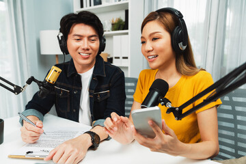 Smiling radio influencer hosts wearing headphones with channel social media online live streaming with talking script note with phone and laptop to listeners at morning at studio record. Infobahn.