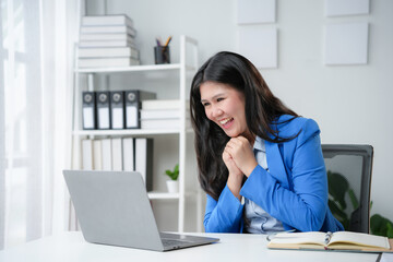 Obraz premium Asian businesswoman is working in her bright office, looking excitedly at her laptop screen and holding her hands together in joy