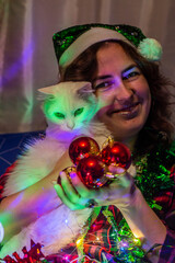 A girl in a New Year's hat holds red New Year's toys and a fluffy white cat in her hands.