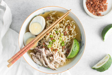 Overhead view of noodles in clear broth with hard boiled eggs, shredded chicken and a lime wedge, top view of ramen noodles in chicken broth on a marble countertop