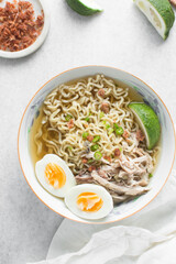 Overhead view of noodles in clear broth with soft boiled eggs, shredded chicken and a lime wedge, top view of ramen noodles in chicken broth on a marble countertop