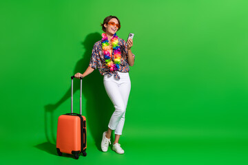Full size photo of satisfied girl wear print shirt colorful necklace hold bag order taxi on smartphone isolated on green color background