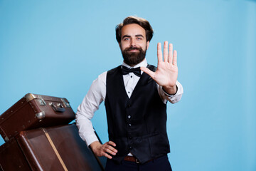 Employee showing stop sign on camera, hotel concierge in suit and tie implying refusal and rejection with hand raised in studio. Professional bellboy standing against blue background.