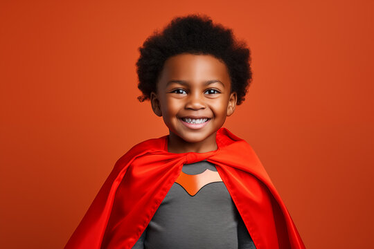 Cute young African American super hero boy in a red cape on a bright orange background smiling at the camera