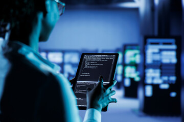 Computer operator builds firewall systems in server room housing advanced infrastructure. Facility manager runs code on tablet to upgrade data center rigs handling demanding workloads