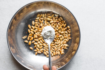 Overhead view of roasted peanuts being coated with egg and flour in a silver bowl, flatlay of roasted peeled peanuts being battered in egg and flour, process of making coated peanuts or cracker nuts