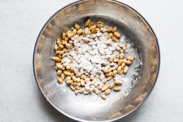 Overhead view of roasted peanuts being coated with egg and flour in a silver bowl, flatlay of roasted peeled peanuts being battered in egg and flour, process of making coated peanuts or cracker nuts