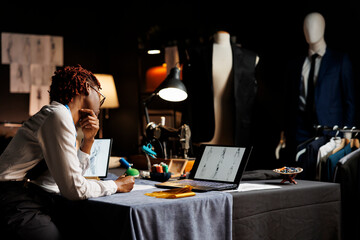 Professional seamstress handdrawing dress designs for upcoming fashion runway show, looking online for design inspirations. Couturer in tailoring studio sketching bespoken sartorial stylish collection