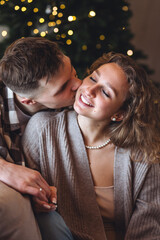 Close up portrait of beautiful loving couple spending time together on Christmas holidays, embracing, kissing. Cozy home interior, atmospheric light, decorated new year fir tree on background