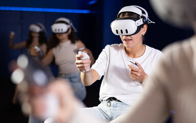 Young woman sitting on stool and enjoying virtual reality game with VR headset and controllers in...