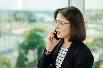 A pretty woman in glasses is standing by the window in a transparent office. A woman rests looking out the window in the office