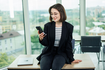 Woman business manager smiling working on laptop in office. An employee works in an office with large windows.