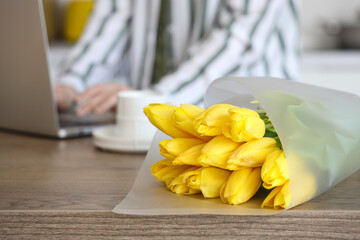 Woman using modern laptop at table with bouquet of yellow tulip flowers