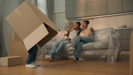 Happy family relocation moving day loan mortgage African American mother father on sofa playing child little boy son jump from box play hide and seek game kid jumping hug parents have fun home weekend