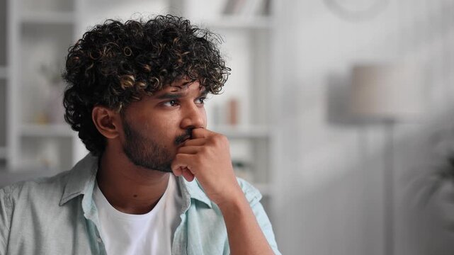 Sad upset Indian man sitting in his living room, thinking about the burdens of life and problems at work. Tired guy, burnout at work, emotional exhaustion.