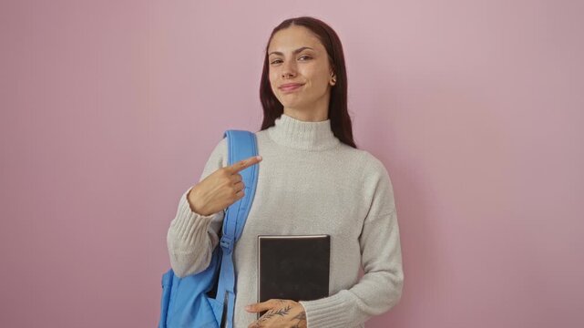 Young hispanic woman student in backpack holding book happily pointing with finger and hand over isolated pink background, smiling and excited