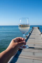 Hand with glass of cold rose wine from Provence and wooden yacht boota pier on white sandy beach Plage de Pampelonne near Saint-Tropez, summer vacation in France