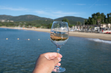 Hand with glass of cold rose wine from Provence and wooden yacht boota pier on white sandy beach Plage de Pampelonne near Saint-Tropez, summer vacation in France
