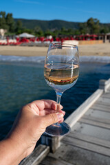 Hand with glass of cold rose wine from Provence and wooden yacht boota pier on white sandy beach Plage de Pampelonne near Saint-Tropez, summer vacation in France